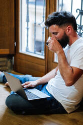 Bearded man sipping hot drink and reading data from laptop while sitting on floor and working on remote project at home - ADSF10980