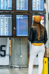 Rückansicht einer gesichtslosen Dame in modischem Outfit mit Gepäck, die in der Nähe des Zeitplans steht und die Abflugzeit auf der digitalen Anzeige überprüft, während sie auf den Flug im Flughafenterminal wartet - ADSF10983