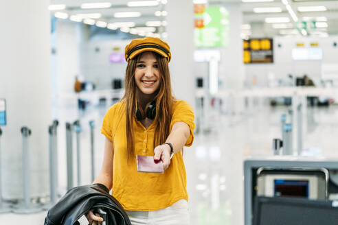 Fröhlich lächelnde Dame in bunten stilvollen Kleidern und Kopfhörer geben Pass und Blick auf die Kamera beim Stehen am Check-in-Schalter im Flughafen-Terminal - ADSF10984