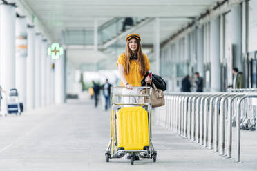 Lächelnde Dame in Freizeitkleidung und Kopfhörern mit Lederjacke, Tasche und Reisepass in der Hand schaut in die Kamera und trägt einen Gepäckwagen in der Nähe eines modernen Flughafengebäudes - ADSF10986