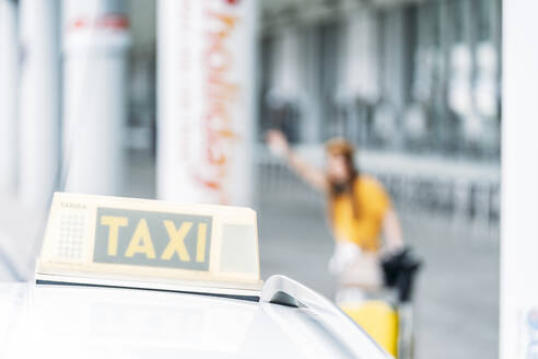 Selektiver Fokus einer anonymen Frau in gelber Kleidung, die ein Taxi ruft, während sie vor einem unscharfen Flughafengebäude steht und die Hand hochhebt - ADSF10988