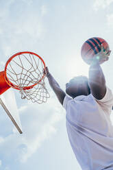 Kräftiger, energiegeladener afroamerikanischer Sportler, der sich auf dem Spielplatz an einen Basketballkorb hängt, nachdem er den Ball im Netz versenkt hat - ADSF10991