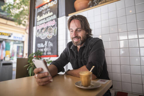 Mann genießt in einem Café, während er auf sein Mobiltelefon schaut - CAVF88265