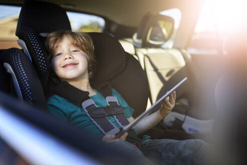 Porträt eines lächelnden Jungen, der ein digitales Tablet benutzt, während er auf dem Rücksitz eines Autos sitzt - CAVF88289