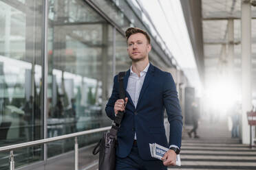 Businessman with newspaper and bag looking away while walking in city - DIGF12852