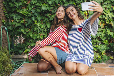 Smiling sisters while taking selfie on smart phone while kneeling in backyard - DLTSF01081