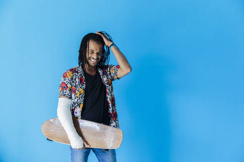 Smiling man with fractured arm holding skateboard against blue background - MRAF00576