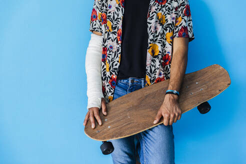 Man with fractured arm holding skateboard while standing against blue background - MRAF00577