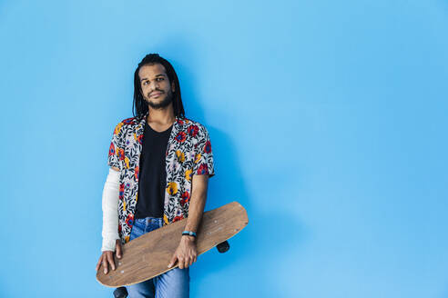 Mittlerer erwachsener Mann mit gebrochenem Arm, der ein Skateboard vor blauem Hintergrund hält - MRAF00578