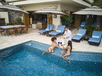 Female friends toasting drinks while relaxing at poolside in holiday villa - ABAF02310