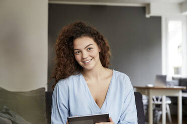 Smiling beautiful young woman with long brown curly hair holding digital tablet at home - FMKF06250