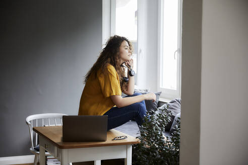 Nachdenkliche Frau, die durch das Fenster schaut, während sie eine Pause von der Arbeit im Wohnzimmer im Büro zu Hause macht - FMKF06255