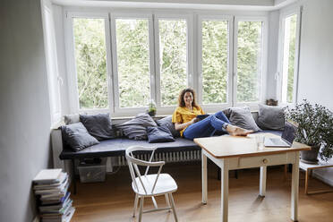 Woman with digital tablet relaxing on sofa against window at home - FMKF06261