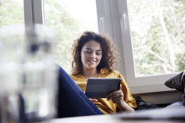 Frau benutzt digitales Tablet, während sie zu Hause am Fenster sitzt - FMKF06263