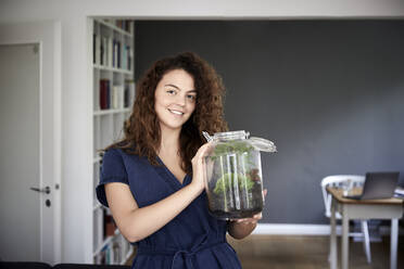 Smiling young woman holding terrarium jar at home - FMKF06273