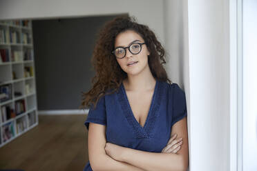 Beautiful woman wearing eyeglasses while leaning on wall with arms crossed at home - FMKF06277