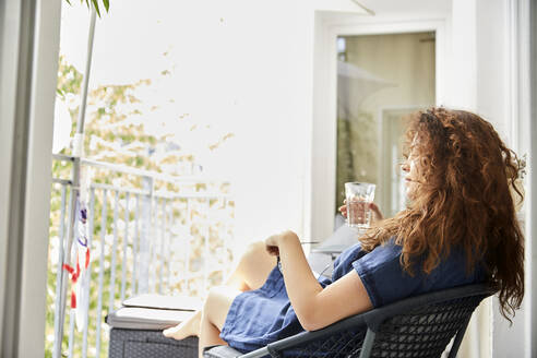 Frau hält ein Glas in der Hand und sitzt auf einem Stuhl auf dem Balkon zu Hause - FMKF06279