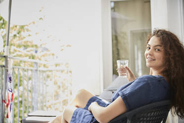 Junge Frau hält ein Glas und entspannt sich auf einem Stuhl auf dem Balkon - FMKF06280