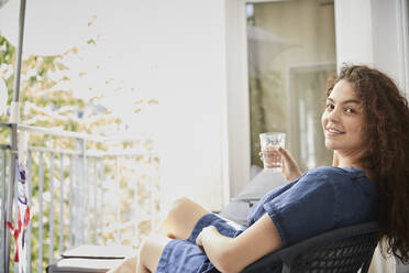 Young woman holding glass while relaxing on chair at balcony - FMKF06280