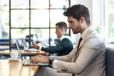 Serious businessman using laptop at table in cafe - JSRF01032