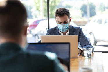 Businessman wearing protective face mask while using laptop in cafe during coronavirus outbreak - JSRF01038