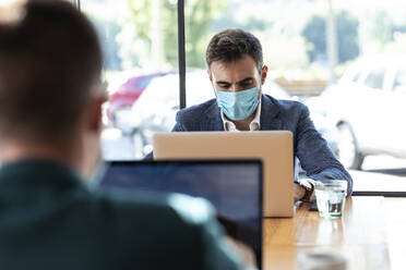 Geschäftsmann trägt während des Ausbruchs des Coronavirus einen Mundschutz, während er in einem Café einen Laptop benutzt - JSRF01038