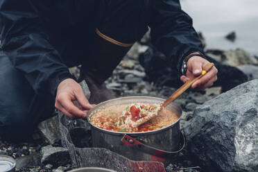 Ein Wildcamper rührt in einem Topf, der über einem Feuer kocht, heißes Essen um. - MINF15061
