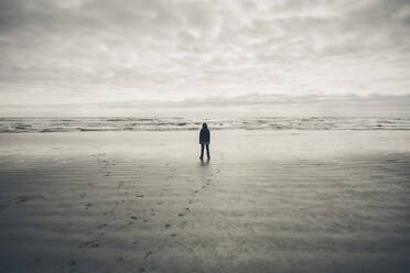 Teenage boy standing on vast beach waves and overcast sky in distance - MINF15067