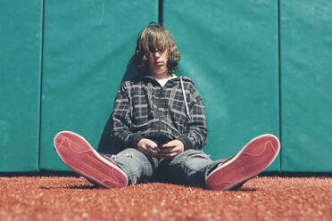 Teenage boy sitting against padded wall at sports field holding mobile phone - MINF15070