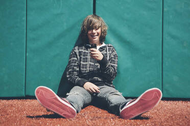 Teenage boy sitting against padded wall at sports field holding mobile phone - MINF15071