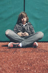 Teenage boy sitting against padded wall at sports field holding mobile phone - MINF15072