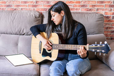 Gitarre spielende Frau, die zu Hause auf ihrer Couch sitzt und mit Online-Lektionen mit einem digitalen Tablet mit einem digitalen Tablet mit einem leeren Bildschirm von oben lernt - ADSF10992