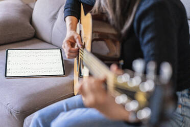 Woman playing guitar sitting on her couch at home and learning with online lessons with a digital tablet with a digital tablet with a blank screen from above - ADSF10993