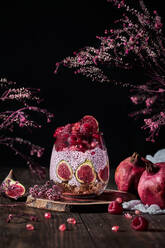 Chia-Pudding mit frischen Feigen und Himbeeren mit Granatapfel und Sahne im Glas am Tisch zwischen rosa Blütensträußen auf schwarzem Hintergrund - ADSF11010