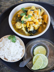 Von oben Teller mit köstlichem vegetarischem Curry und Limettenscheiben auf einem Tablett auf Holzuntergrund neben Tellern mit Reis und frischen Limetten - ADSF11019