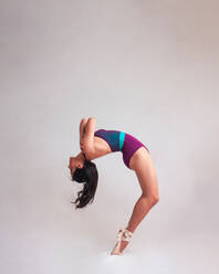 Side view of graceful woman in leotard and pointe shoes bending back while dancing against gray background - ADSF11043