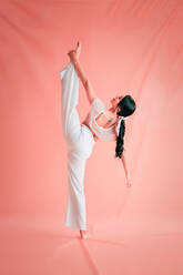 Back view of slim female in white outfit doing splits while dancing gracefully on pink background - ADSF11044