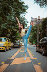 Full body cheerful female in casual clothes doing splits with raised arm and smiling while dancing on asphalt road amidst cars on busy street of modern city - ADSF11046