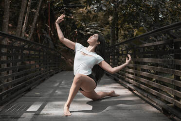 Full body barefoot female with outstretched arms and closed eyes dancing on wooden bridge on sunny day in peaceful garden - ADSF11047