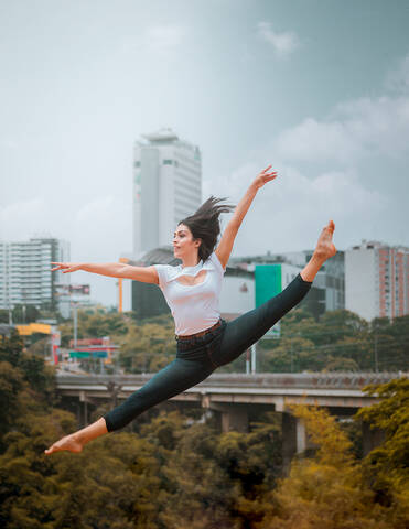 Full body energetic barefoot woman in casual clothes jumping and doing splits while dancing against contemporary city and cloudy sky stock photo