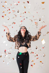 Slim woman in stylish outfit smiling and looking up while trying to catch golden confetti during party against gray background - ADSF11050