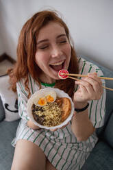 From above cheerful redhead female laughing and picking noodles from bowl of tasty ramen while sitting on couch at home - ADSF11056