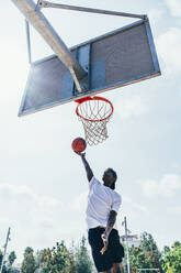 Kräftiger, energiegeladener afroamerikanischer Sportler, der sich auf dem Spielplatz an einen Basketballkorb hängt, nachdem er den Ball im Netz versenkt hat - ADSF11139