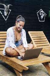 Fröhliche Frau in Freizeitkleidung, die mit gekreuzten Beinen auf einem hölzernen Liegestuhl auf einer tropischen Terrasse sitzt und im Laptop surft - ADSF11187