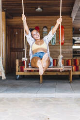 Gebräunte glückliche Frau in Freizeitkleidung, die auf einer Schaukel sitzt und über eine Holzwand in Costa Rica hinwegschaut - ADSF11189