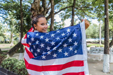 Seitenansicht einer fröhlichen Frau, die in die Kamera blickt und eine amerikanische Flagge wie einen Umhang trägt, während sie an grünen Bäumen in Costa Rica entlanggeht - ADSF11193