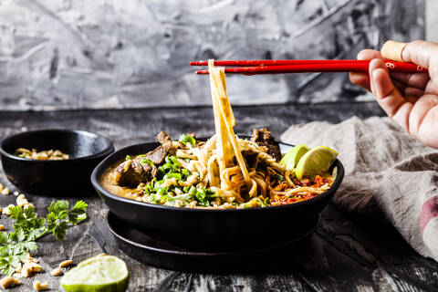 Hand of woman eating bowl of red curry soup with rice noodles, beef, vegetables, scallion, peanuts and lime stock photo