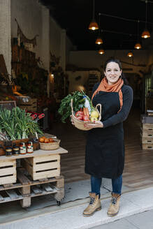 Smiling saleswoman holding vegetable basket while standing at store entrance - RDGF00057