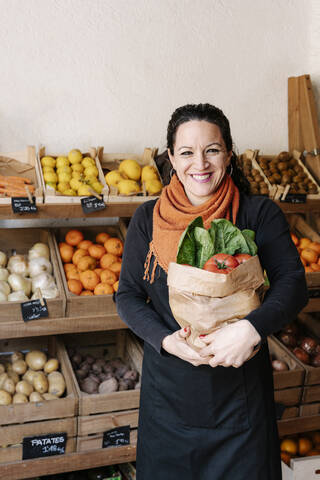 Cheerful saleswoman holding bag of vegetables standing against boxes at store stock photo