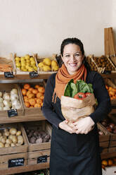 Cheerful saleswoman holding bag of vegetables standing against boxes at store - RDGF00064